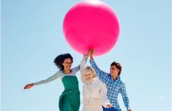 Zwei Frauen und ein Mann halten einen magentafarbenen Ballon, im Hintergrund ist ein klarer blauer Himmel